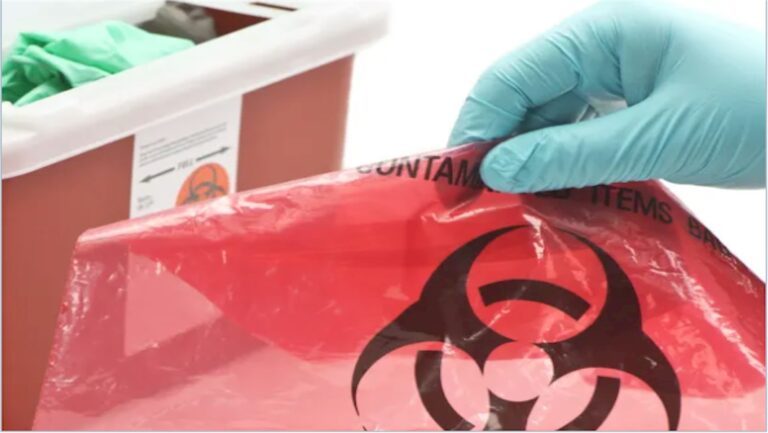 Gloved hand holding a biohazard waste bag beside a sharps container